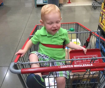 Toddler in green shirt and plaid shorts sits in a Costco shopping cart with eyes partly closed and a funny expression.