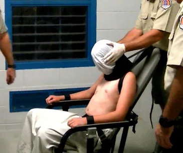 A restrained juvenile detainee in a chair with a hood, flanked by officers, in a detention facility.