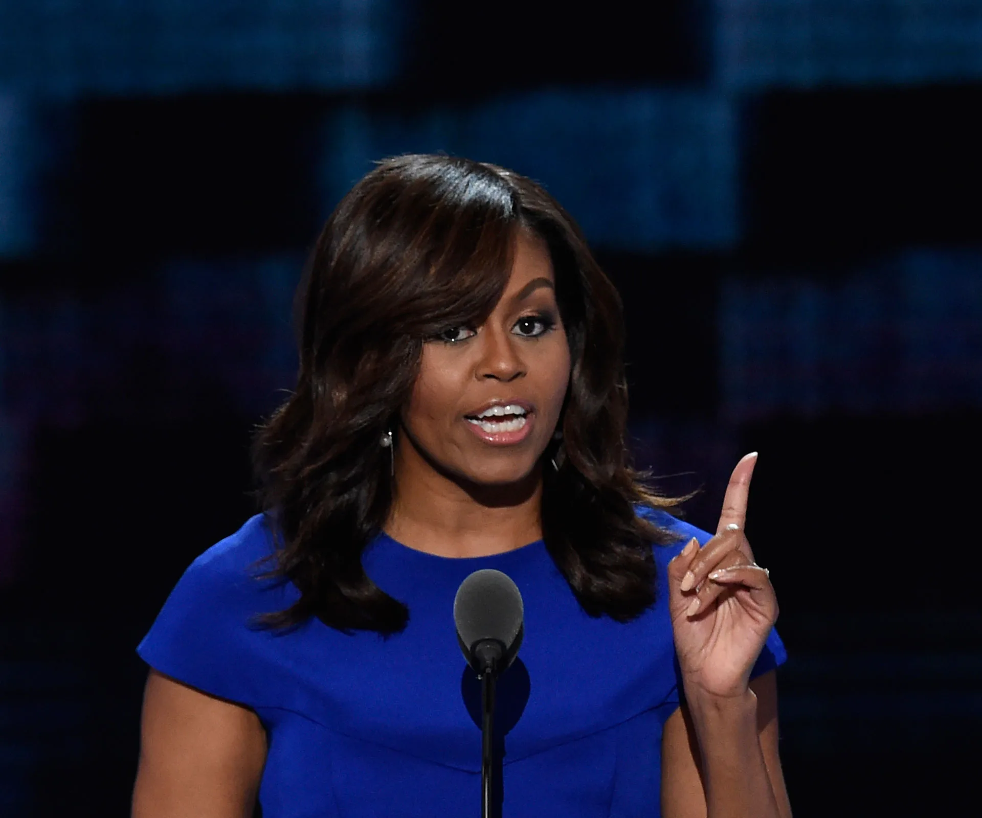 A woman in a blue dress speaks passionately at a podium, gesturing with her right hand.