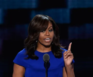 A woman in a blue dress speaks passionately at a podium, gesturing with her right hand.