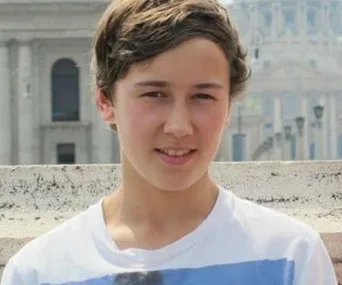 A teenage boy with short brown hair wearing a white T-shirt stands outside with a blurred building in the background.