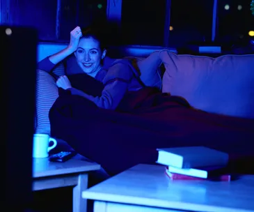 A woman is sitting on a couch, wrapped in a blanket, watching TV with a remote and a mug nearby.