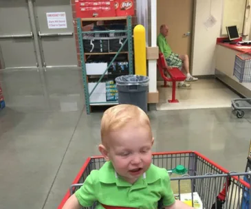 A toddler in a green shirt sits in a shopping cart, with an older man in the background wearing similar colors.