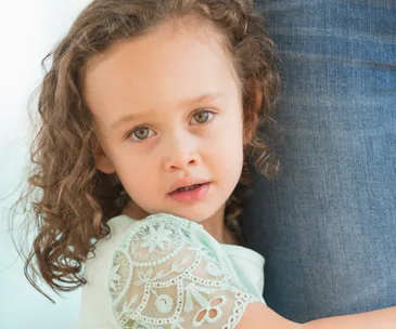 A young girl with curly hair hugs an adult's leg, looking upward with a gentle expression.