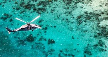 A helicopter flying over a beautiful turquoise ocean with coral reefs visible below.
