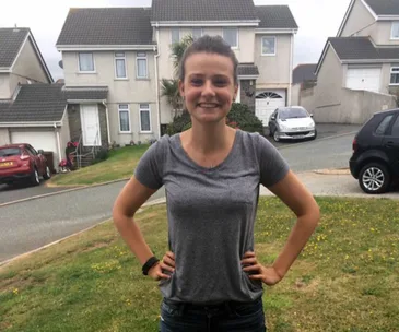 A smiling woman stands confidently with hands on hips in front of suburban houses and parked cars.