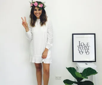 Woman in white dress with floral headband making peace sign, standing beside framed quote.
