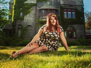 Plus-size woman in a floral dress sits on grass, in front of an ivy-covered stone building, smiling.