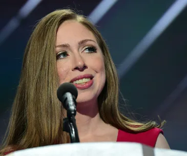 Chelsea Clinton speaking at a podium, wearing a red dress, with a microphone in front of her.