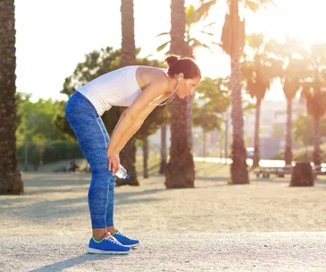 A woman in workout attire rests, bent forward, holding a water bottle, with palm trees and sunlight in the background.