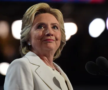 Hillary Clinton speaking at the Democratic National Convention, wearing a white suit, with microphones nearby.