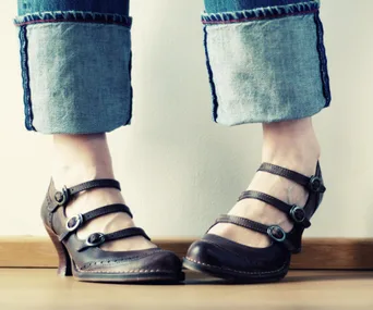 A person wearing brown strappy heeled shoes with buckles and cuffed jeans stands on a wooden floor.