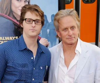 "Two men pose for a photo; one wears a polka dot shirt, the other a light suit, with a movie poster in the background."