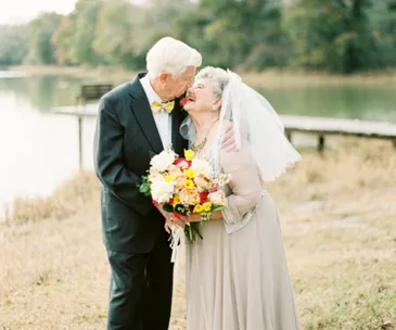 Elderly couple smiling and embracing in wedding attire, holding a bouquet by a lake.