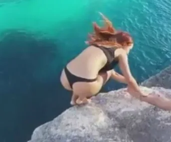 Woman in black swimsuit crouching on cliff edge, reaching for a hand above turquoise water.