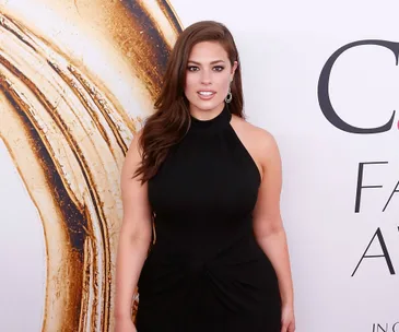 A woman in a black dress poses against a textured wall backdrop at a fashion awards event.