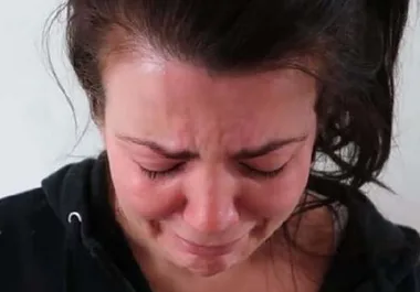 A person with dark hair crying, looking down, wearing a black hoodie against a plain background.