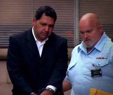 Two men in formal attire, one in handcuffs, walking together outside a building with blinds in the background.