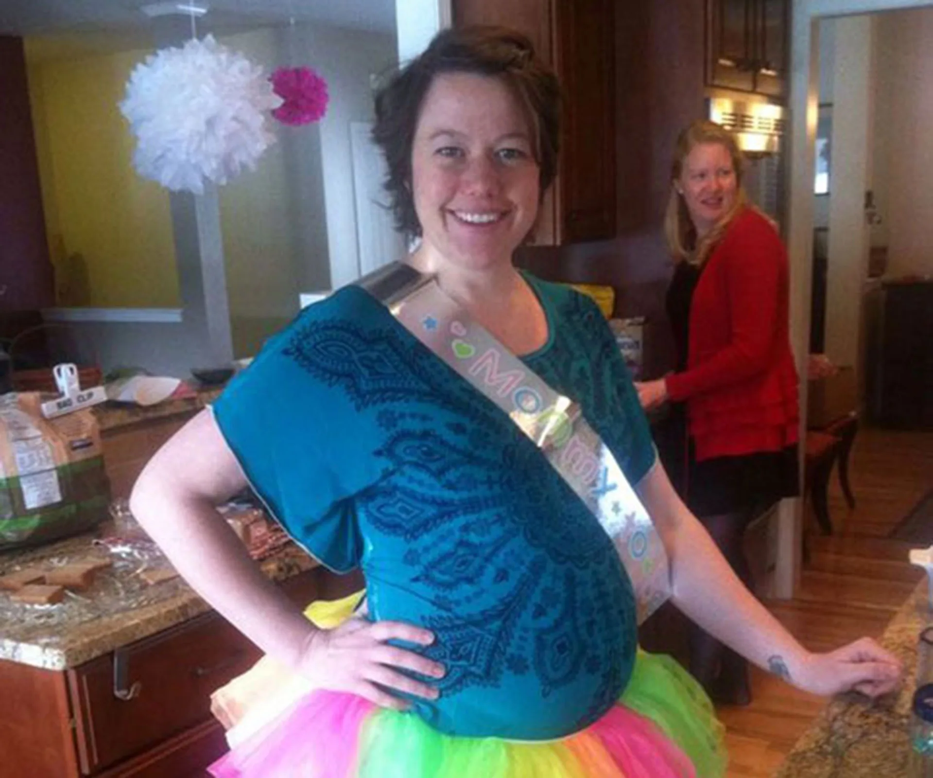 Pregnant woman in a blue top with a "Mommy" sash and colorful tutu, standing in a kitchen with another woman in the background.
