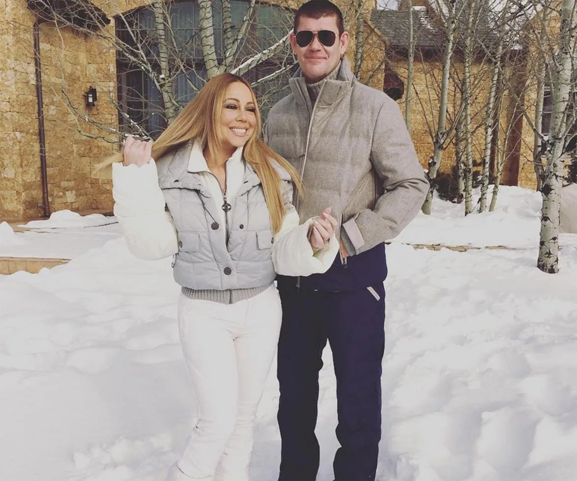 Two people posing in a snowy landscape, wearing winter clothing, with trees and a building in the background.