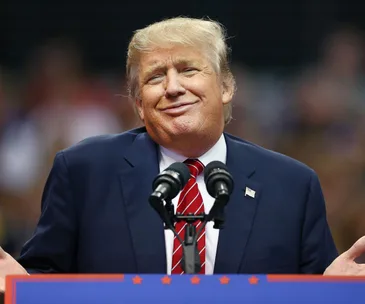 A person at a podium with a microphone, shrugging with a smirk, wearing a suit and a red tie.