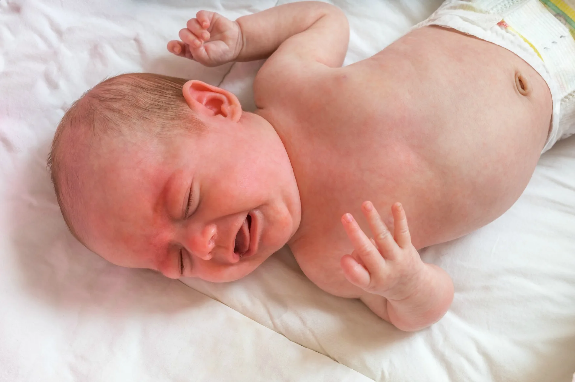 A newborn baby lying on a bed, wearing a diaper, and crying with eyes closed and arms raised.