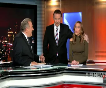 Three news presenters at a studio desk during a live broadcast, one with an arm around another.