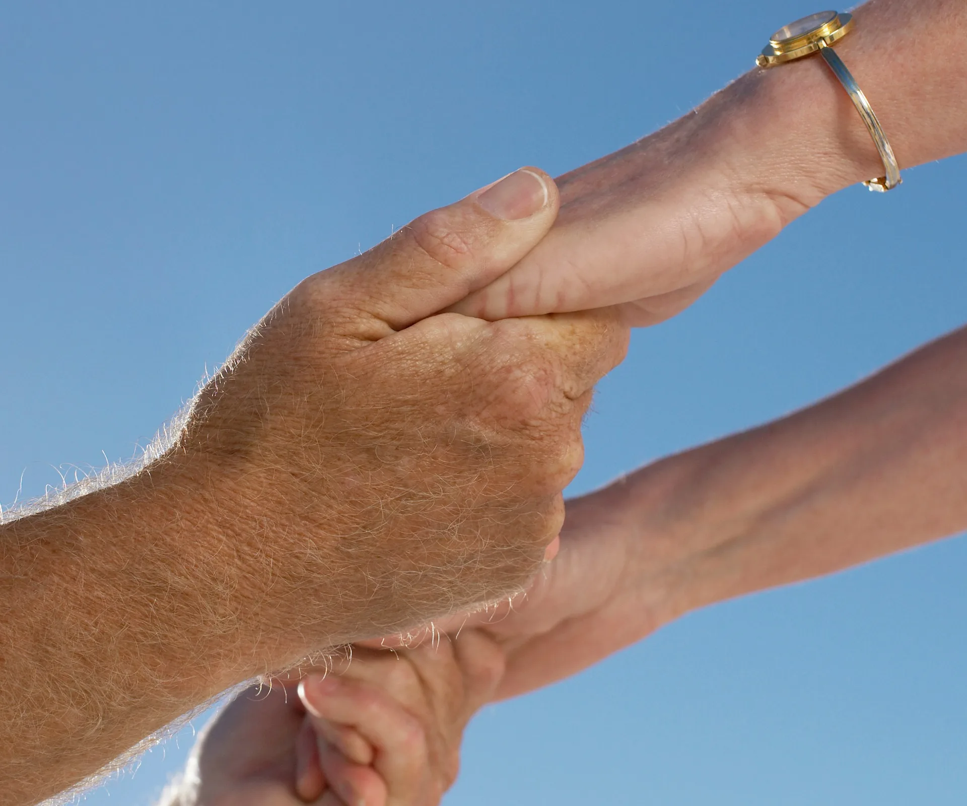 Two elderly hands holding each other against a clear blue sky, one hand wearing a gold watch.