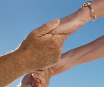 Two elderly hands holding each other against a clear blue sky, one hand wearing a gold watch.