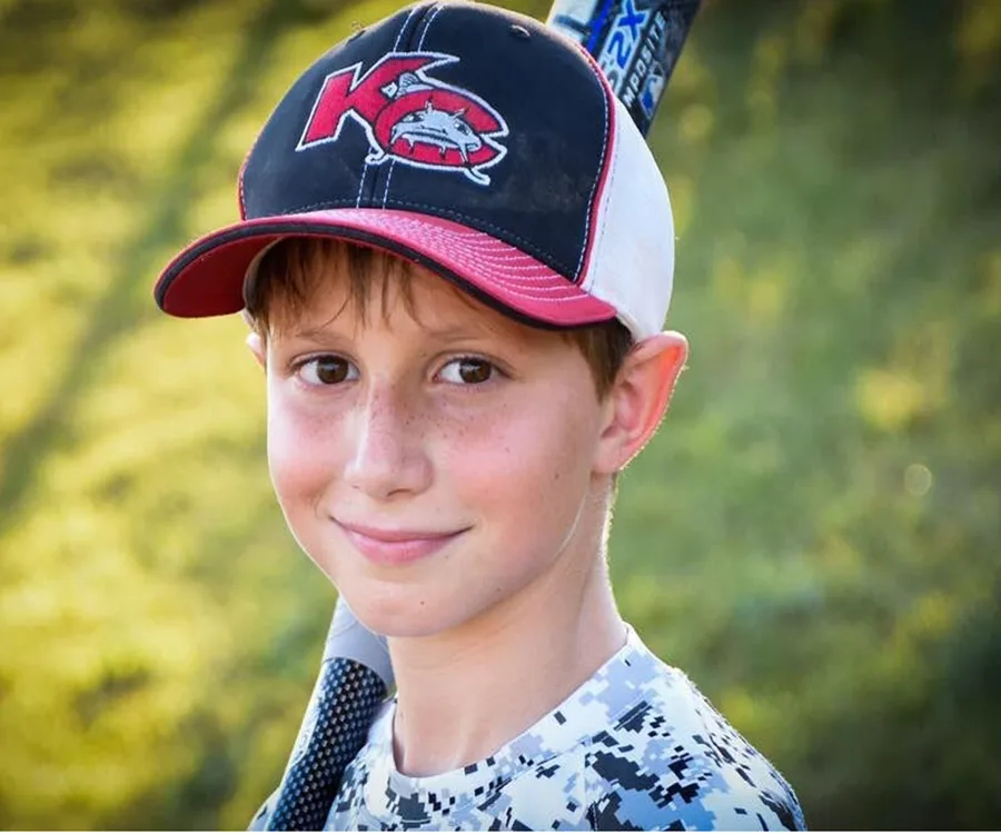 A young boy in a baseball cap and shirt smiles at the camera, holding a baseball bat over his shoulder.