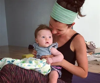 A mother lovingly holds her baby on her lap, both seated on a yoga mat indoors.