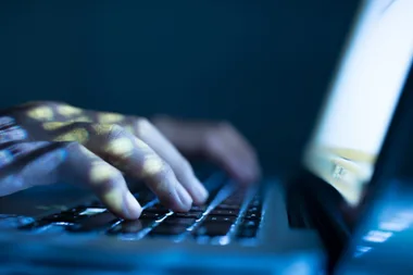 Hands typing on a laptop keyboard in a dimly lit setting with blue digital light reflections.