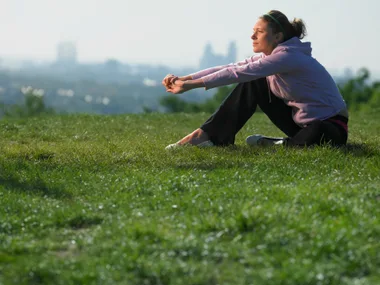 A person sitting on grass, stretching with hands clasped, looking thoughtful, with a blurred cityscape in the background.