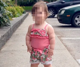 A toddler wearing a pink floral top and camouflage shorts stands in a parking lot.