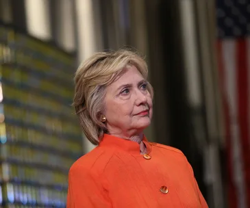 Hillary Clinton in an orange jacket, looking to the side, with an American flag blurred in the background.