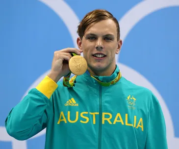 Australian swimmer holds gold medal at Olympics, wearing green Australia jacket, smiling.