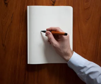 "Person writing on a blank notebook with a pen on a wooden table."