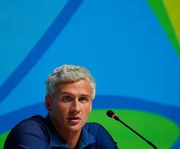 Ryan Lochte speaks at a press conference with a blue and green background, wearing a blue shirt.