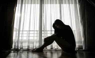A woman sits silhouetted against a window with sheer curtains, head bowed and arms around legs.