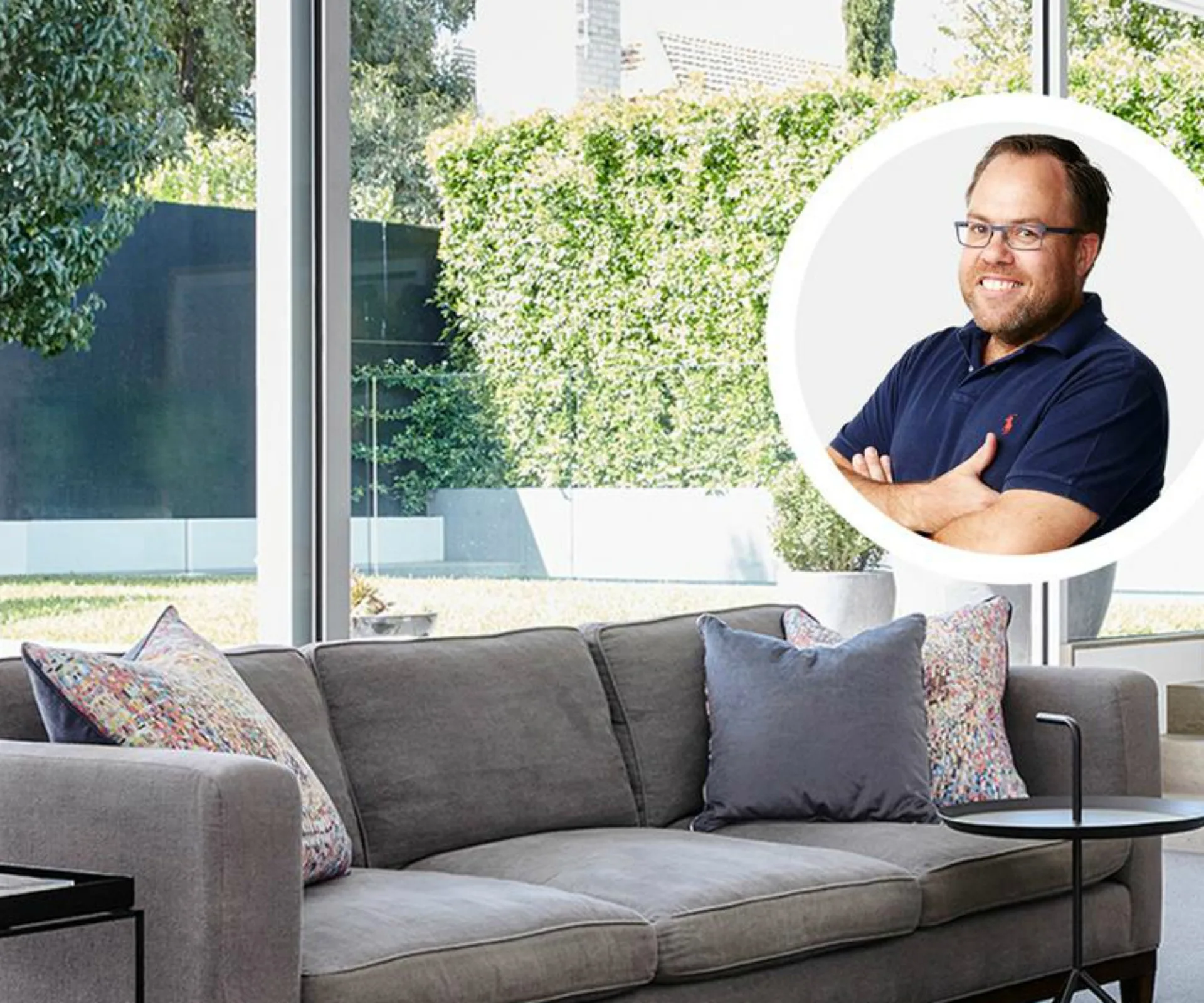 Modern living room with gray couch and colorful pillows, large windows showing greenery; inset smiling man in glasses with arms crossed.