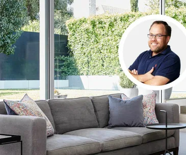 Modern living room with gray couch and colorful pillows, large windows showing greenery; inset smiling man in glasses with arms crossed.