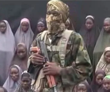 Masked militant with rifle stands in front of a group of seated young girls, some wearing headscarves.