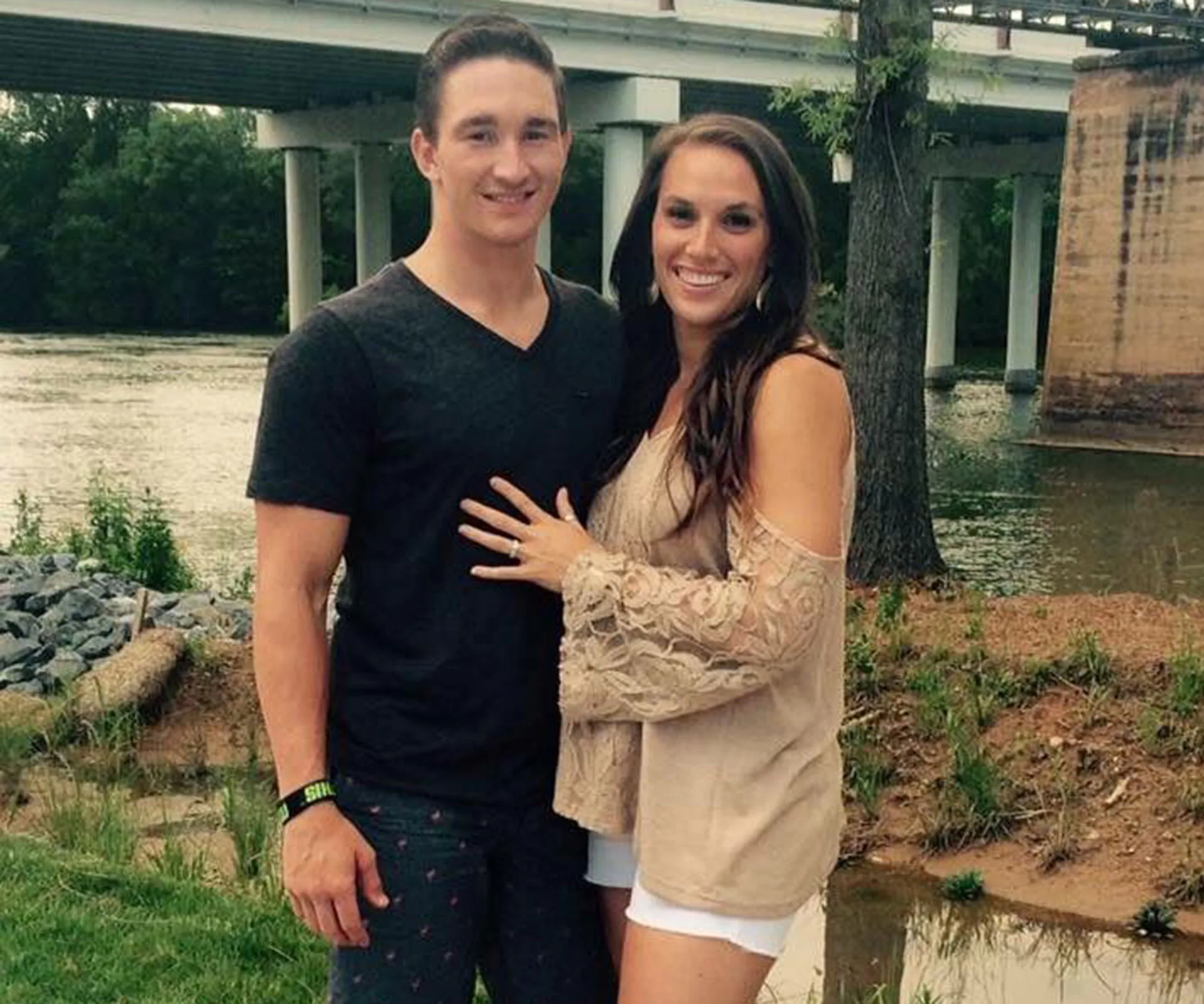 A couple smiling by a river, with trees and a bridge in the background.