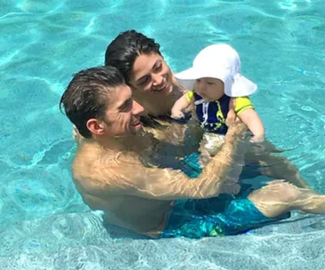 Family enjoying pool time; man holds baby wearing sun hat and swimsuit while woman smiles in background.