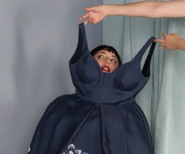 A woman humorously tries on an oversized dress, with another person holding the straps.