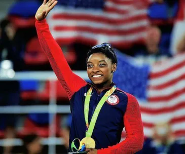 Olympic gymnast smiles with medals at Rio 2016, waving with American flag in background.