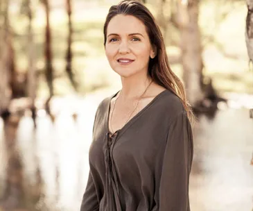 Portrait of a woman with long hair, wearing a dark top, standing outdoors with blurred trees and water in the background.