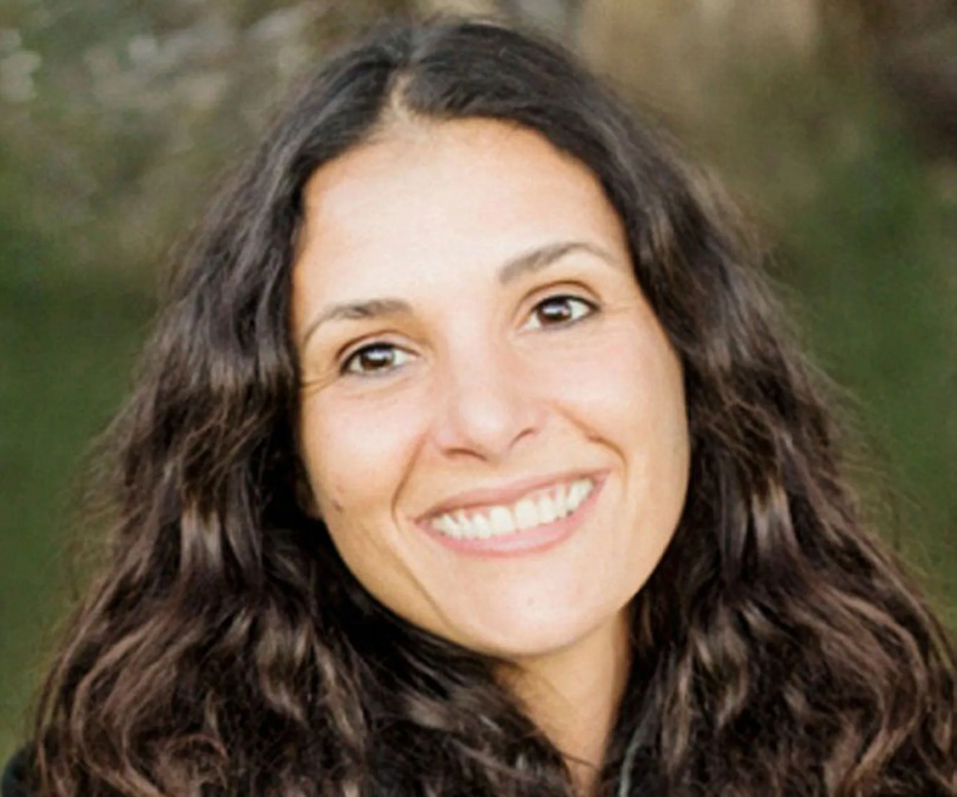 A woman smiling with long curly hair, outdoors with a blurred background.