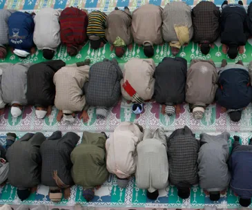 Muslim men in rows performing prayer on patterned mats, seen from above.