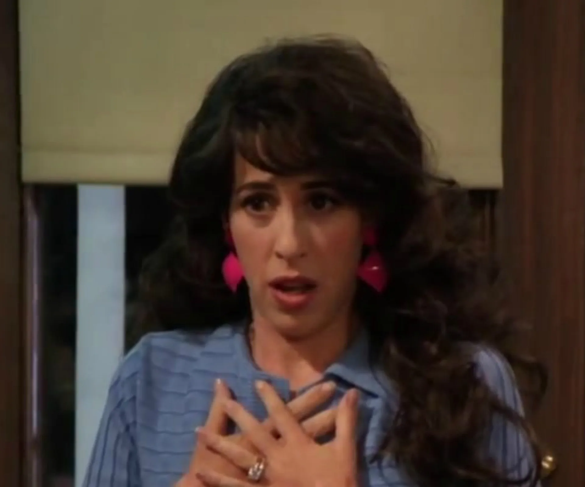 A surprised woman with dark hair and pink earrings, wearing a blue top, holds her hands to her chest.